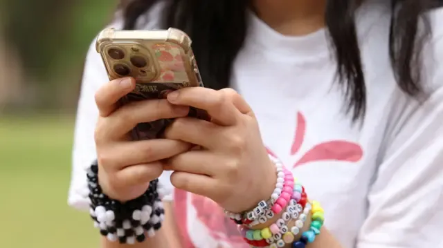 A close-up image of a young girl holding a mobile phone. She wears beaded bracelets on each arm but her face has been cropped out
