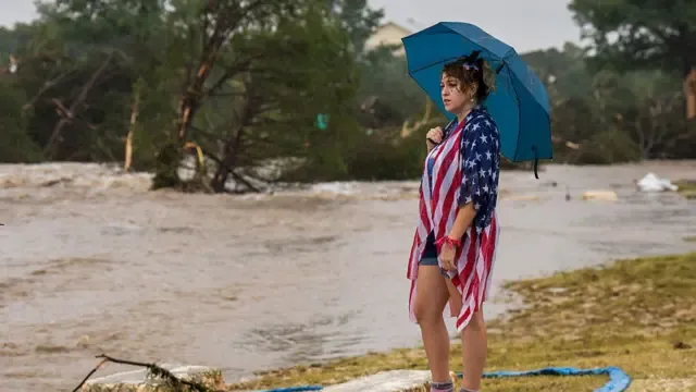 Woman stood on grass looking out at flooded ground, holding umbrella.