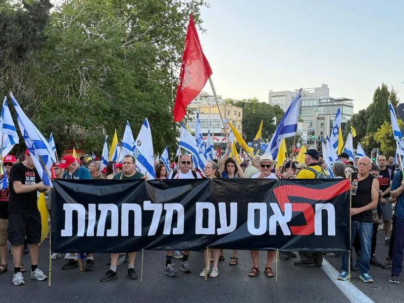 Protesters march along the streets of Haifa, Israel, carrying Israeli flags and yellow flags for the hostages. The sign says "Enough with the wars" following Israel's 12-day conflict with Iran and the war in Gaza, which has surpassed 630 days. The red flag is an anti-government protest symbol.