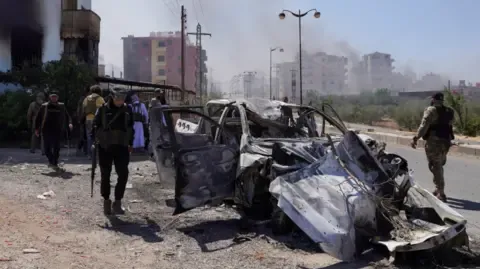 Reuters Bedouin fighters walk near a damaged car in Suweida, following the Syrian presidency's announcement of a ceasefire, 19 July 