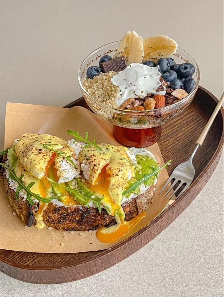 This may contain: an open faced sandwich on a wooden tray next to a bowl of fruit and yogurt