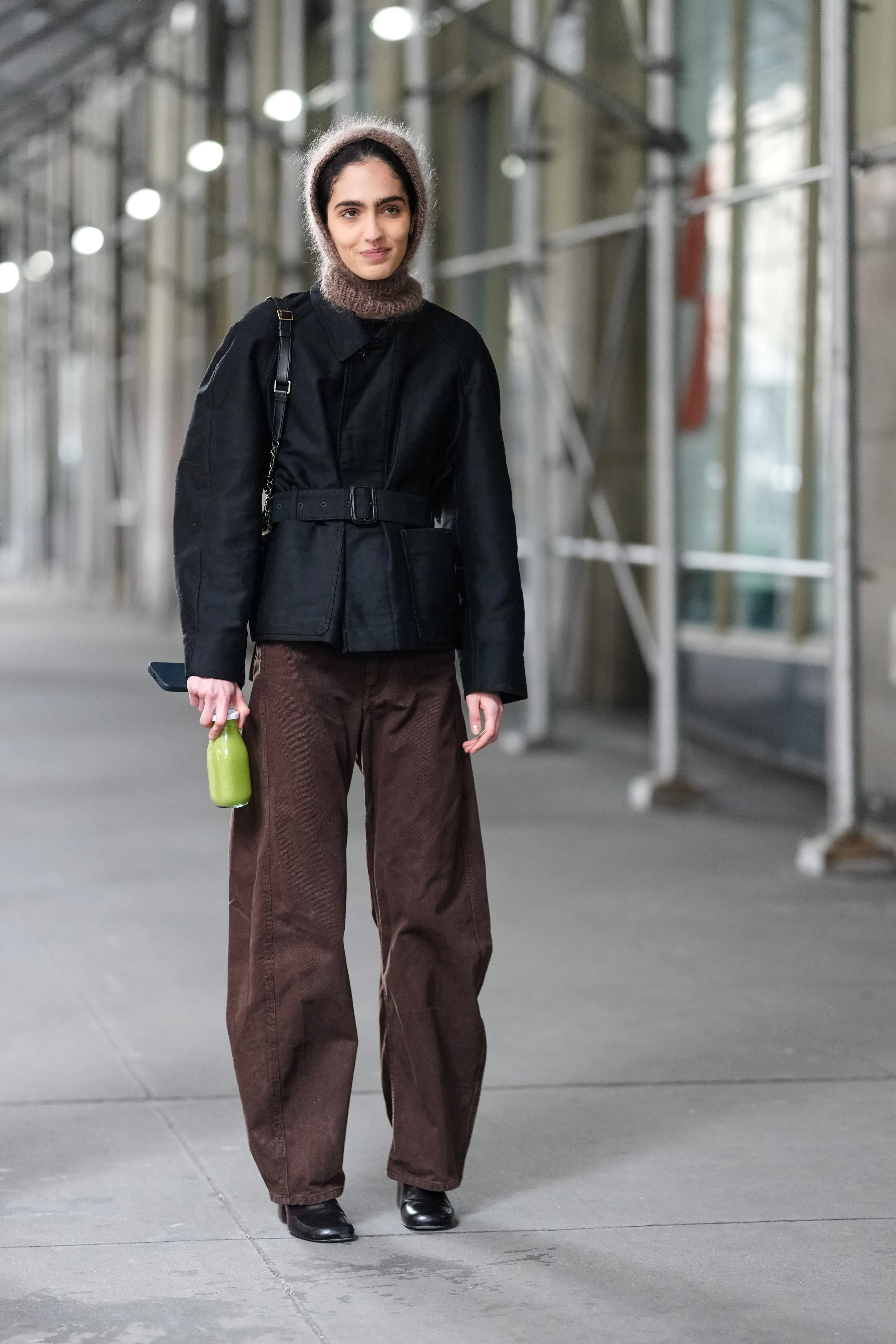 new york, new york february 08: a model wears a brown balaclava hood in wool, a black jacket, brown denim pants / jeans, holds a green bottle of drink, outside altuzarra, during new york fashion week, on february 08, 2025 in new york city. (photo by edward berthelot/getty images)