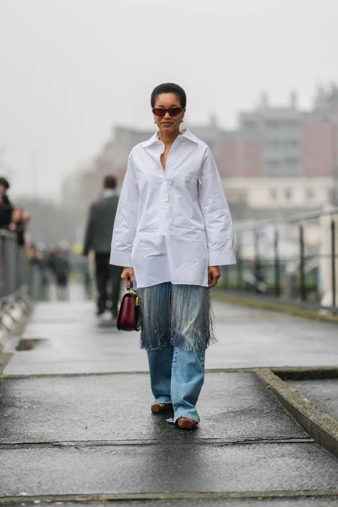milan, italy february 25: tamu mcpherson wears burgundy bordeaux sunglasses, silver earrings, white half buttoned long sleeve oversized shirt, shiny burgundy bordeaux gucci leather bag, light blue denim jean slouchy silver tassel pants, dark brown gucci ballerina suede shoes, outside gucci, during the milan fashion week men&rsquo;s fall/winter 2025 2026 on february 25, 2025 in milan, italy. (photo by edward berthelot/getty images)