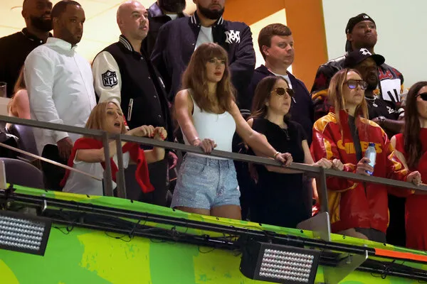 Taylor Swift, in a white tank top and denim shorts, stands against a railing in a luxury box and is surrounded by other people. 