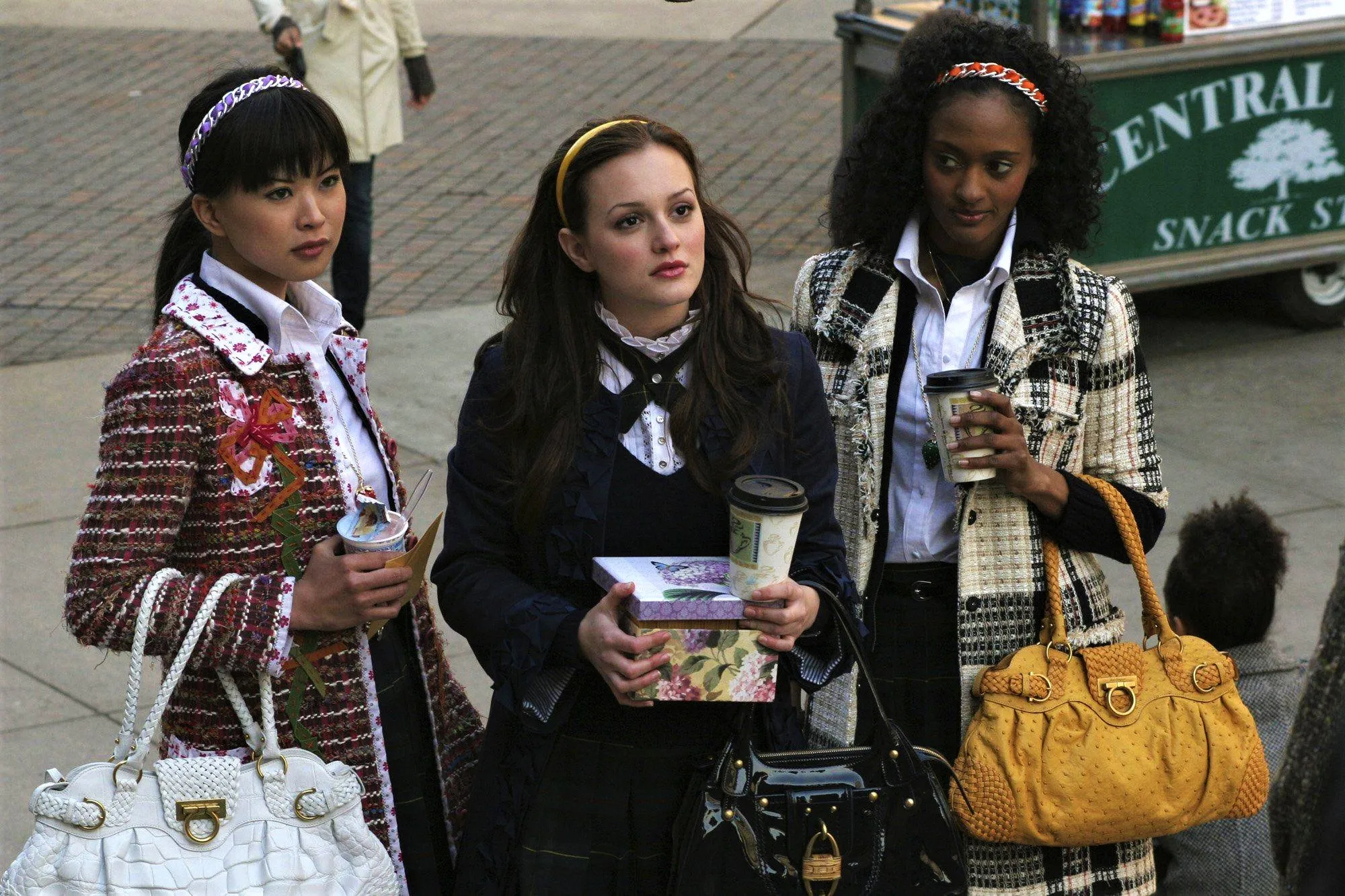 Three girls holding coffees, wearing headbands and coats, and carrying expensive bags pose