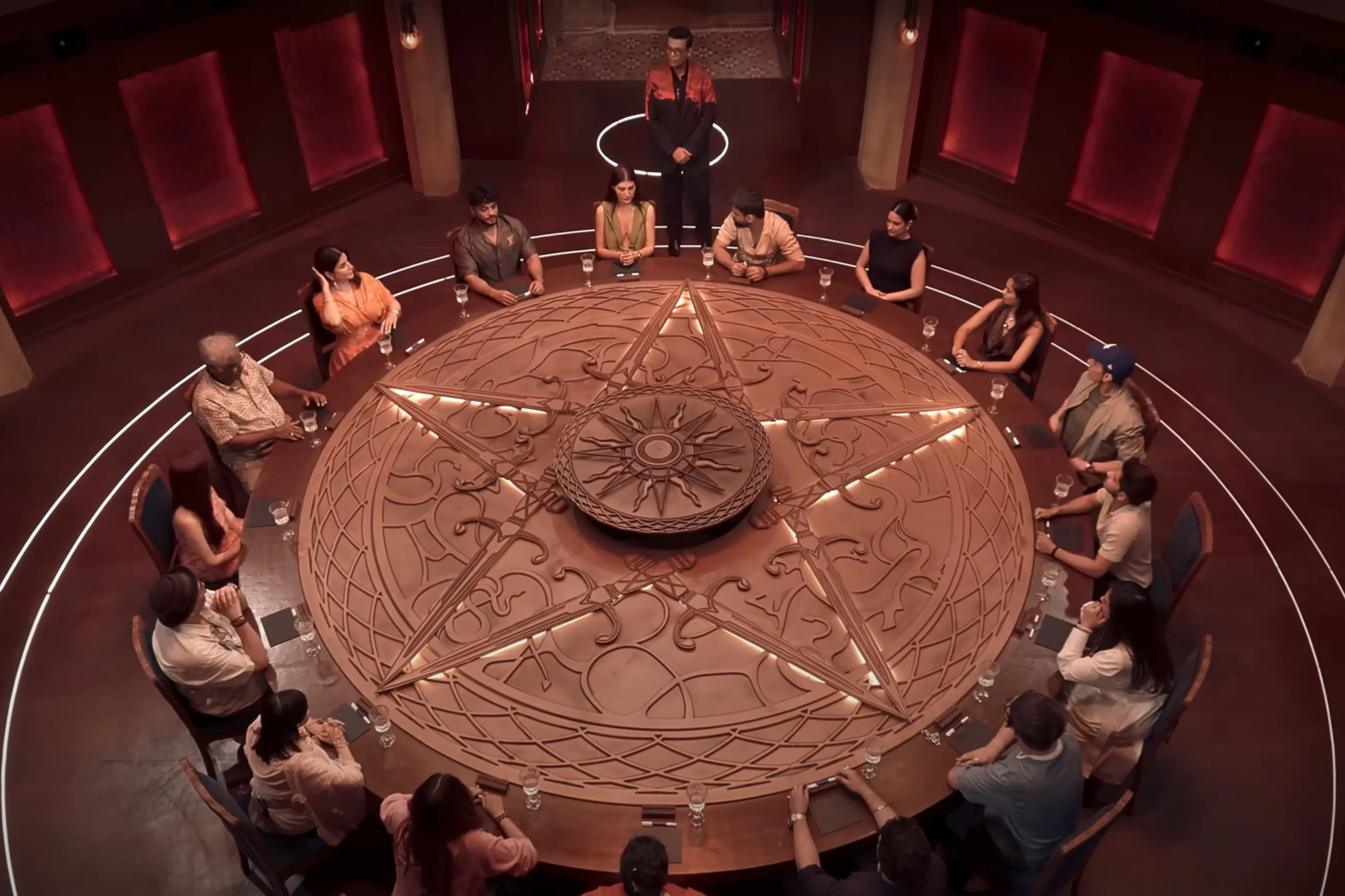 A bunch of people sitting at a round table with a star drawn on it and one person standing at the head of it