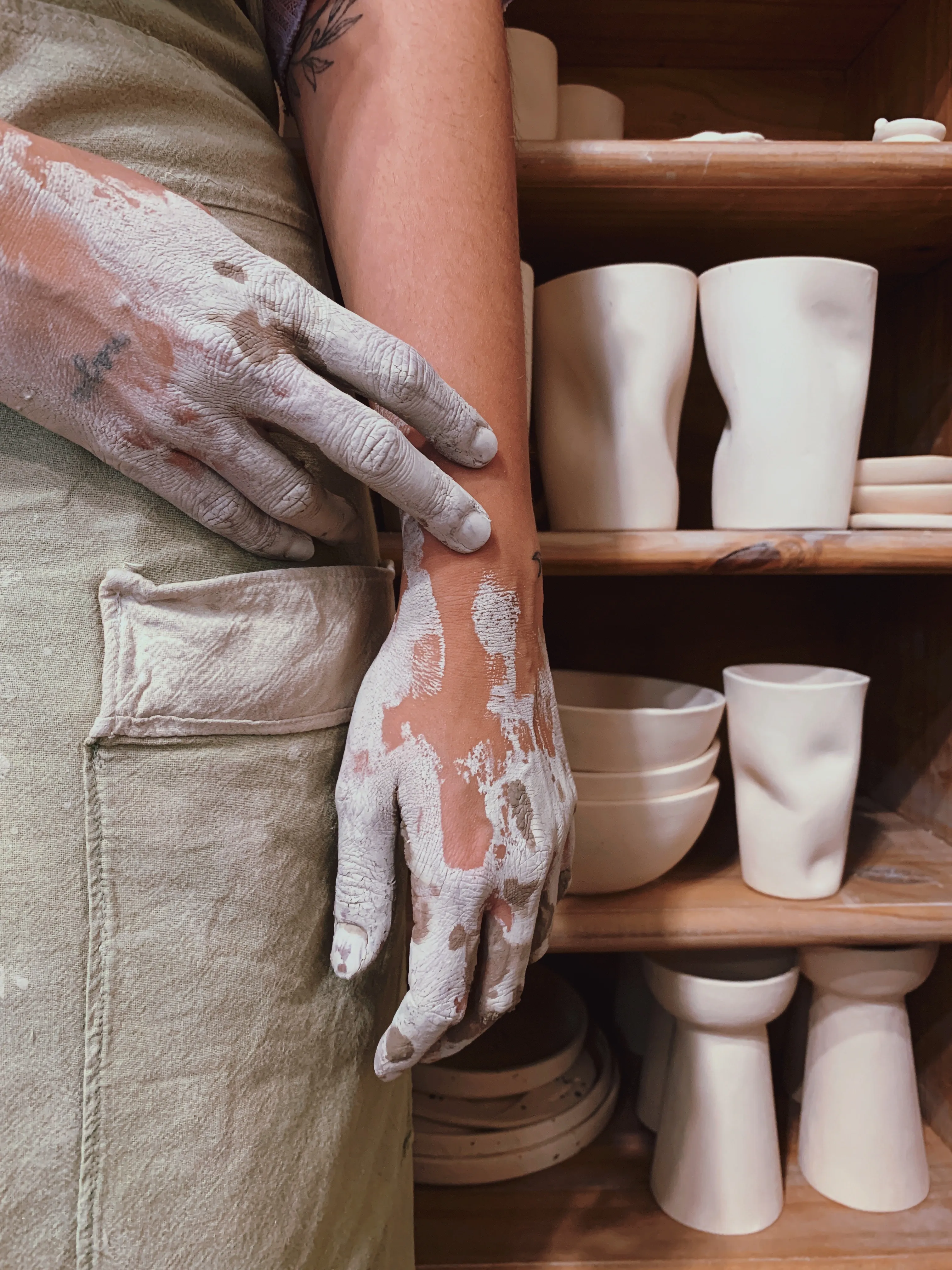 Close-up of a potter’s hand covered in clay with handmade ceramic bowls in the background.