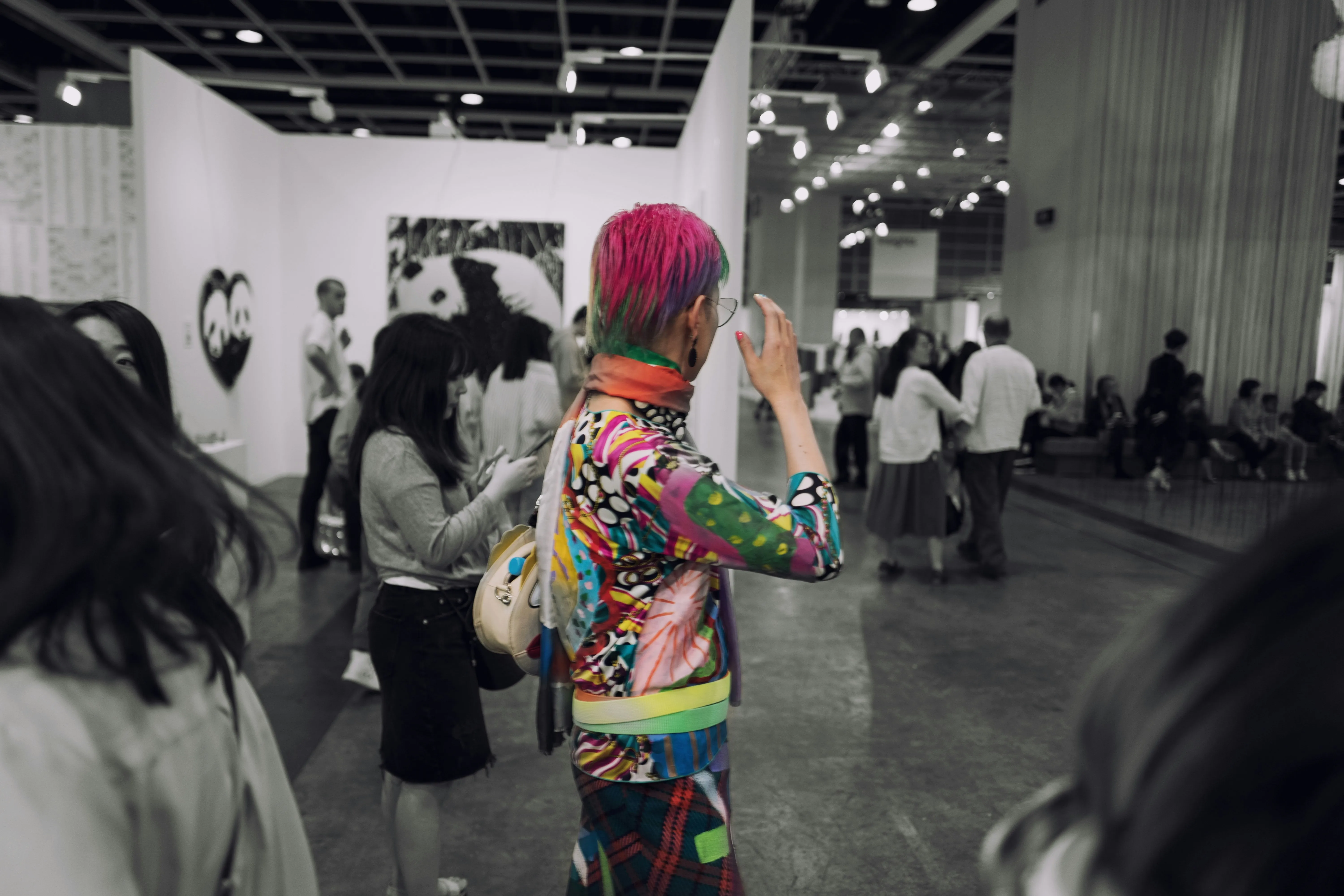 A black and white photograph of a person in colourful clothes and pink hair, depicting queer pride India and drag culture India, in a room full of people that's black and white to show queer safe spaces India and queer visibility India
