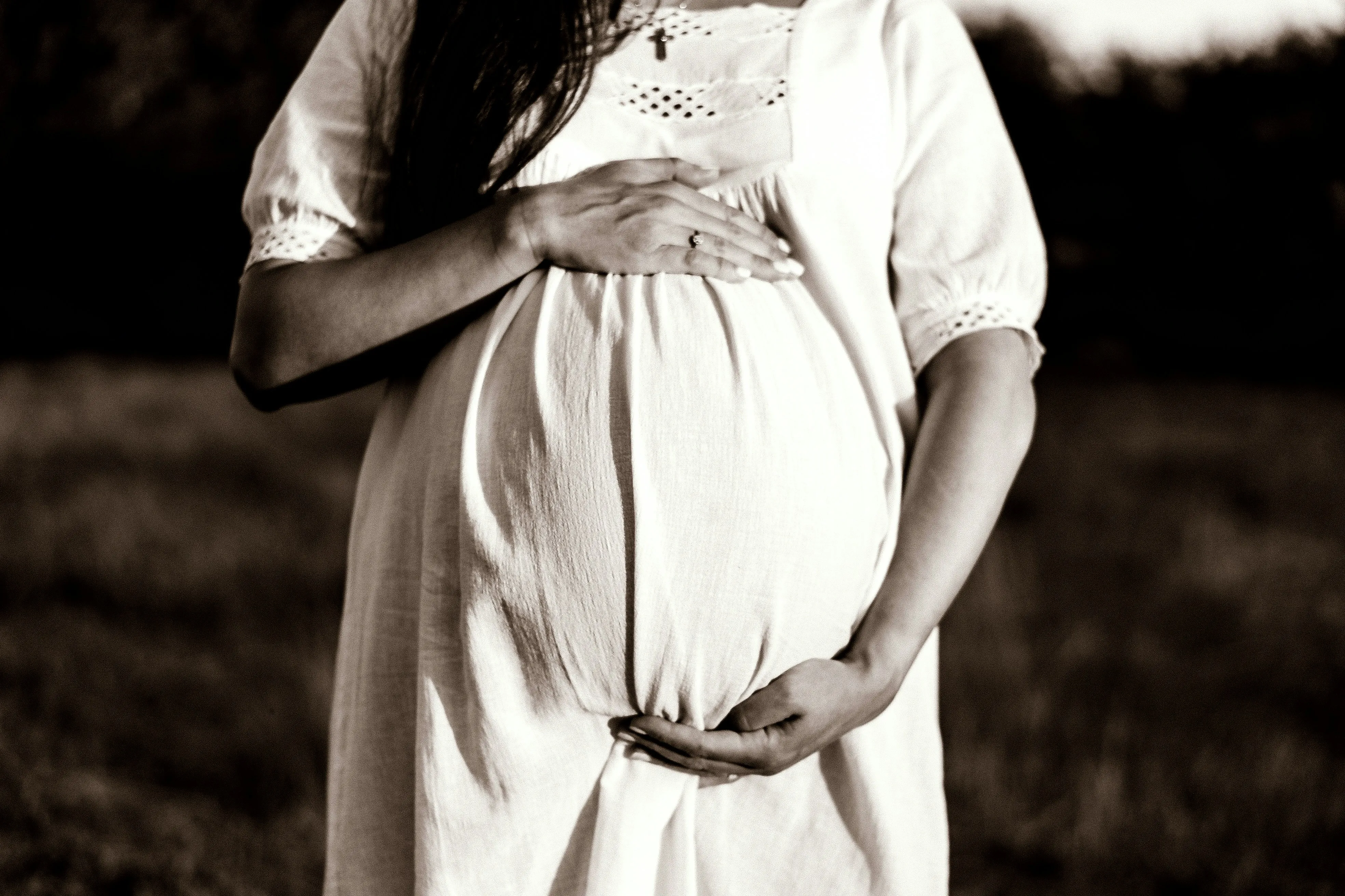 A sepia picture of a woman wearing a white dress holding her baby bump with her arms to show how despite harsh surrogacy laws for queer couples India, queer parenting in India is evolving