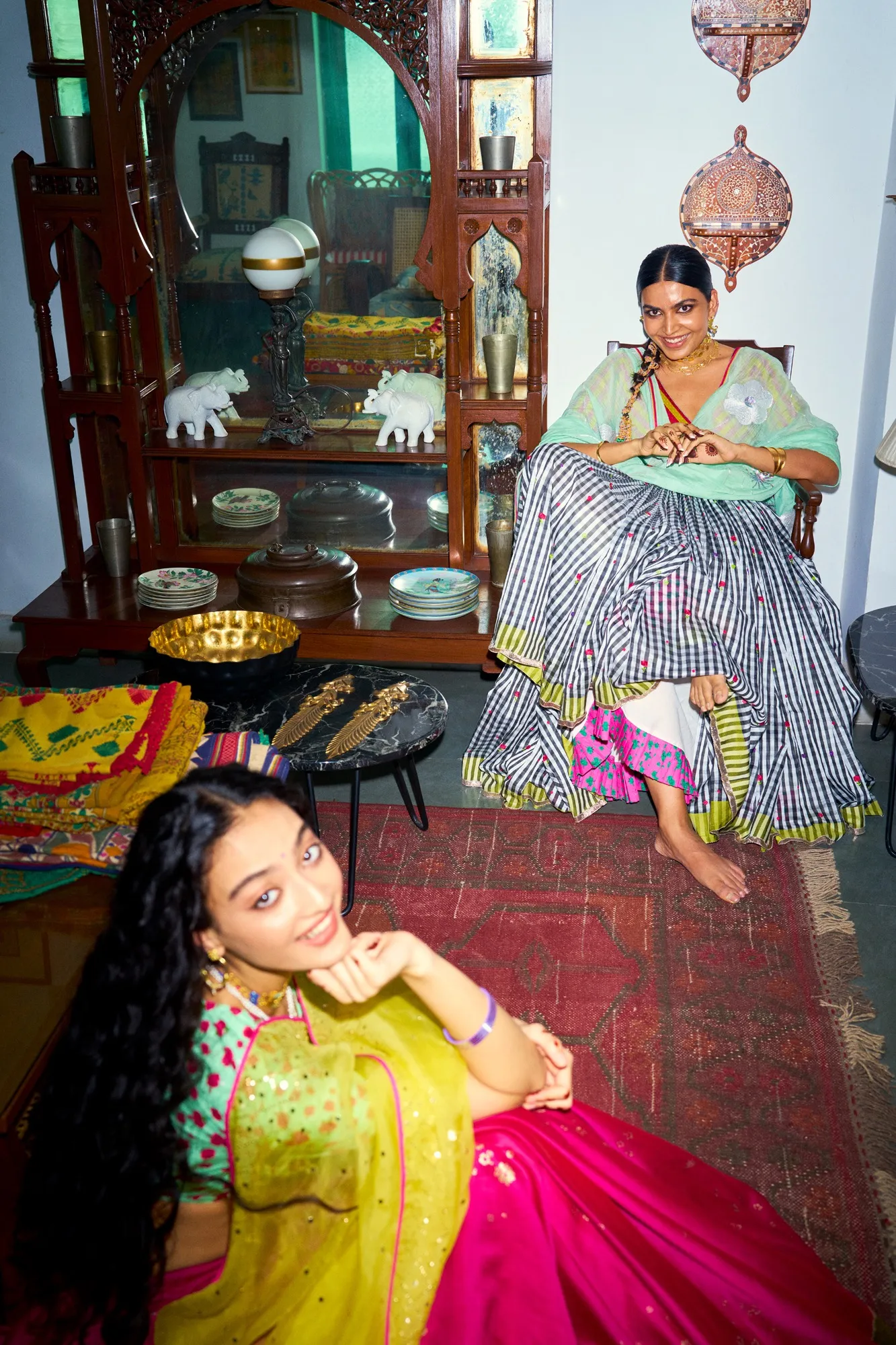Two women wearing Nicobar clothing seated in a colourful, craft-filled home interior with textiles, ceramics and vintage fur