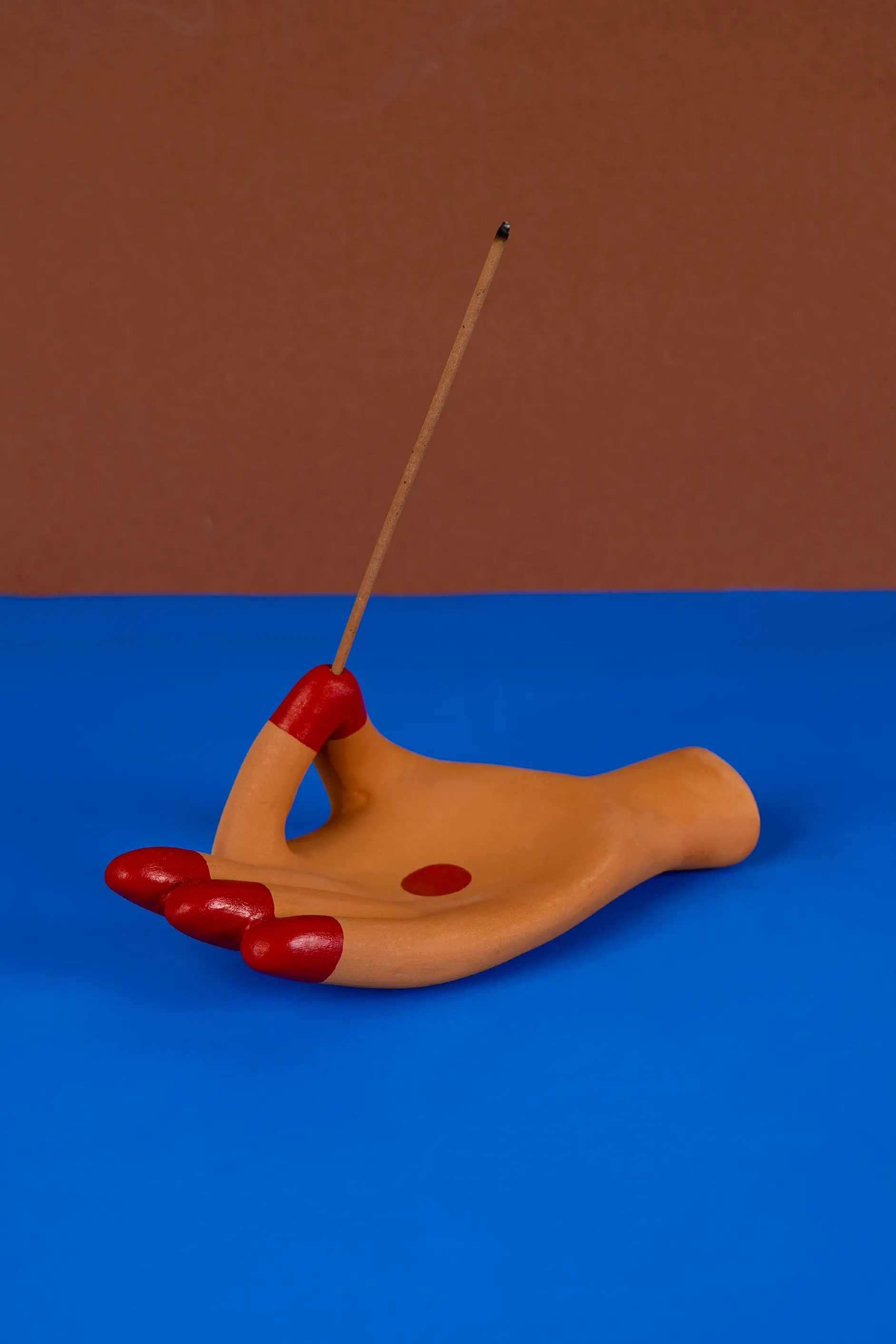 A terracotta-toned hand-shaped incense holder with red painted fingertips holding a lit incense stick against a brown and blue backdrop