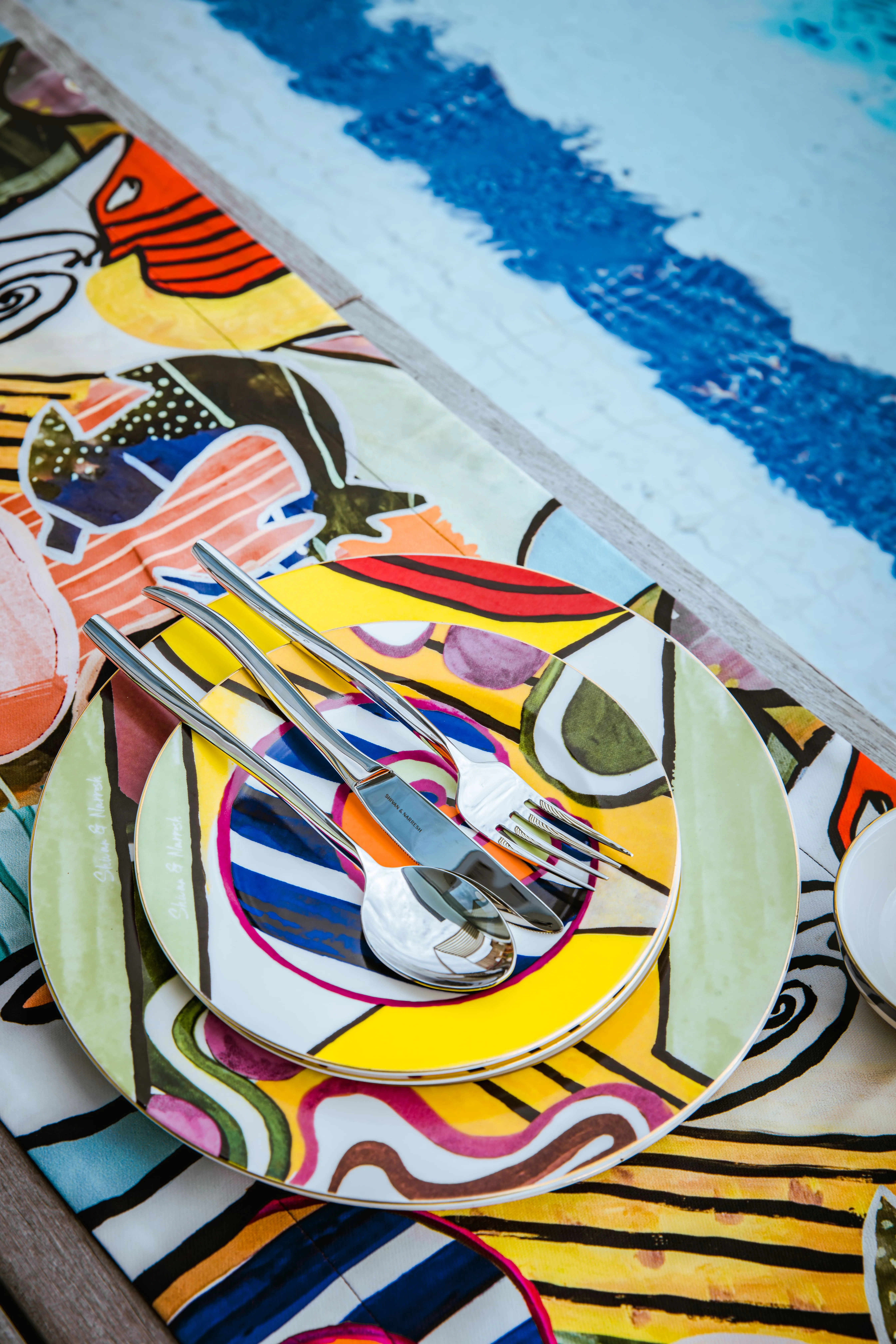Colourful graphic plates and cutlery from Shivan & Narresh arranged on a printed table runner beside a swimming pool.