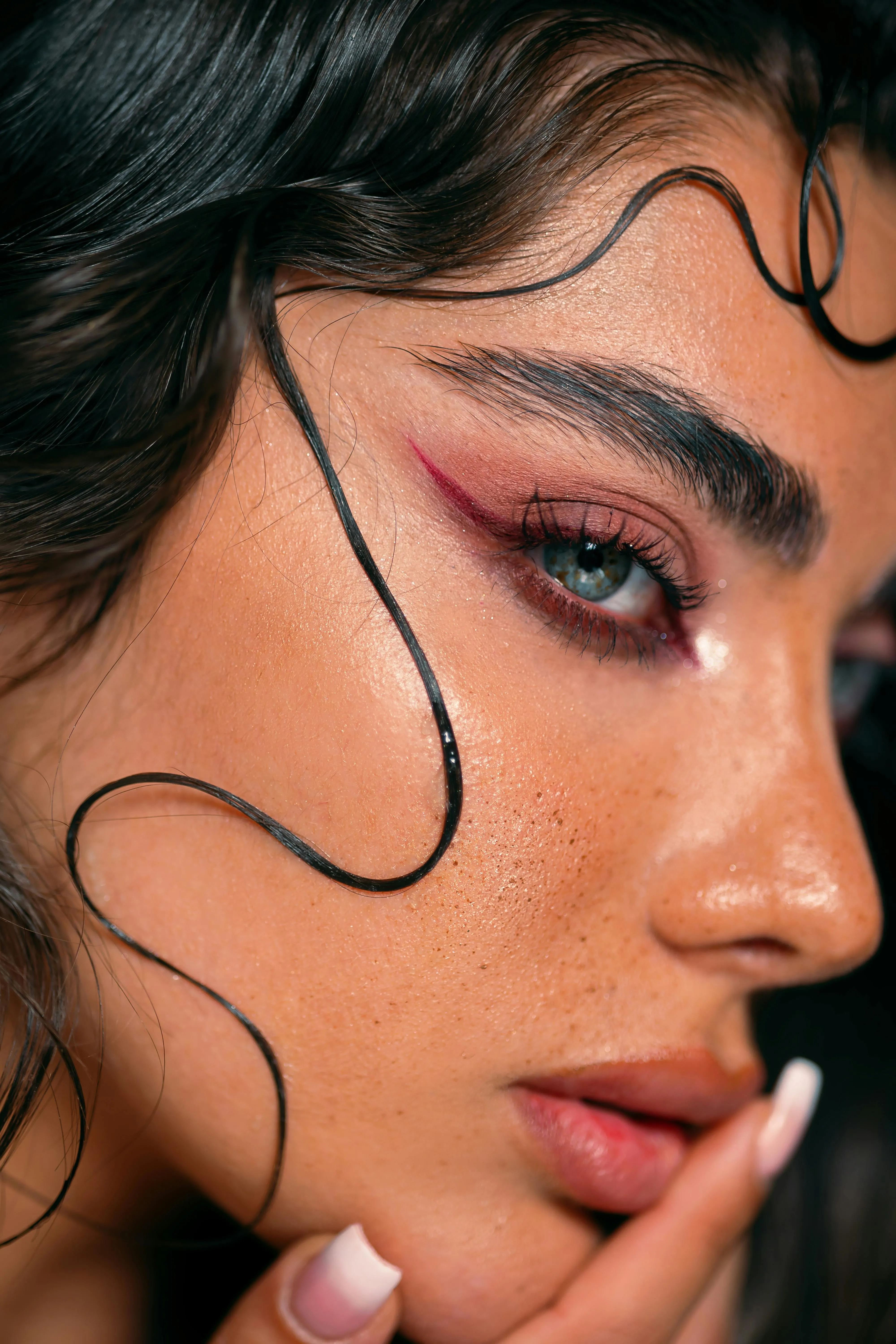 A side profile shot image of a woman with maximalist make-up including a red winged eyeliner, bold brows and wet hair