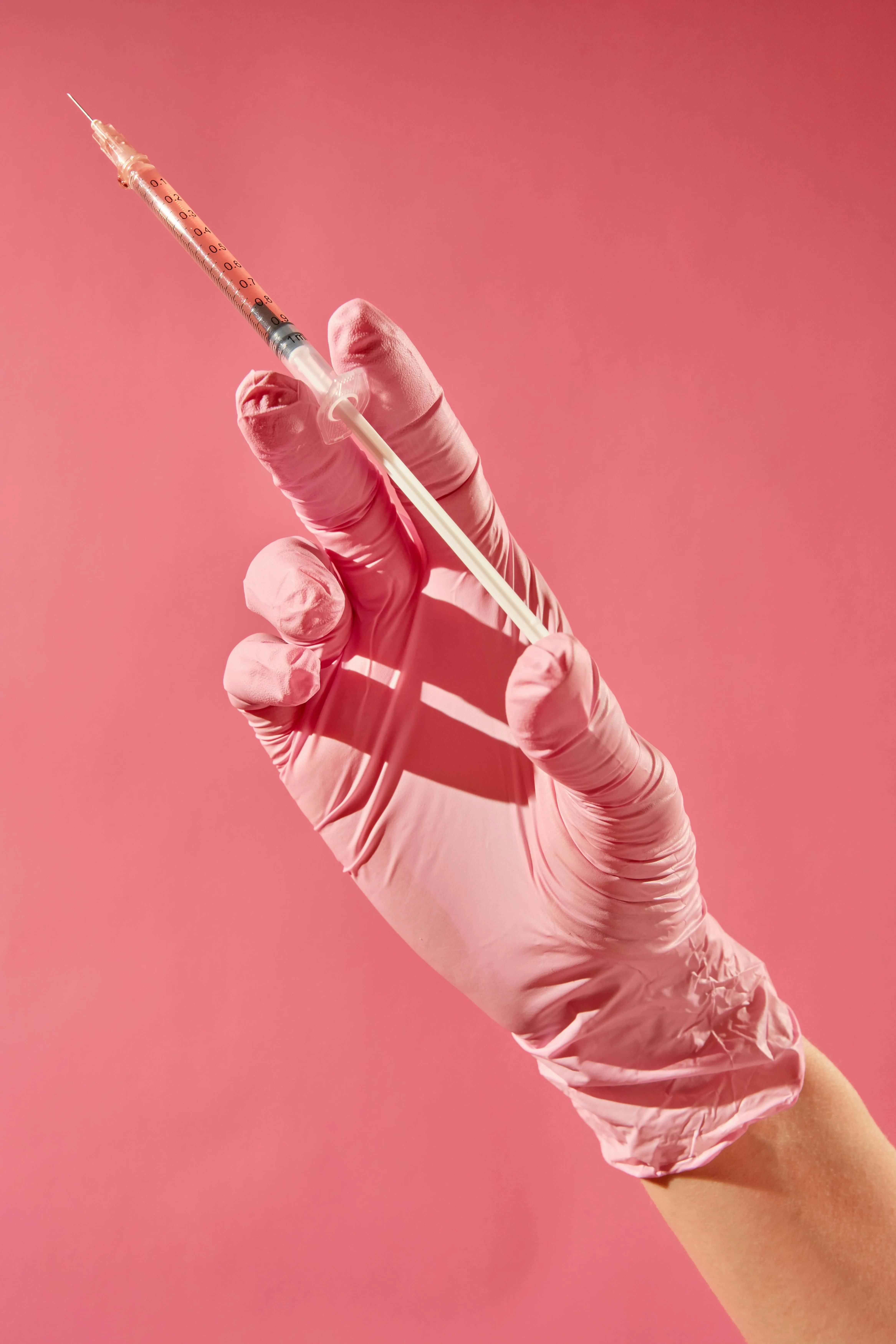 A syringe in a doctor's hand with pink gloves against a pink backdrop