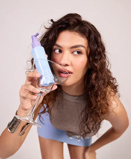 A woman with curly hair with a glass in her hand containing a curly hair product spray