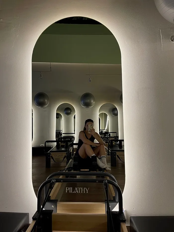A girl taking a mirror selfie on a Pilates reformer in workout clothes