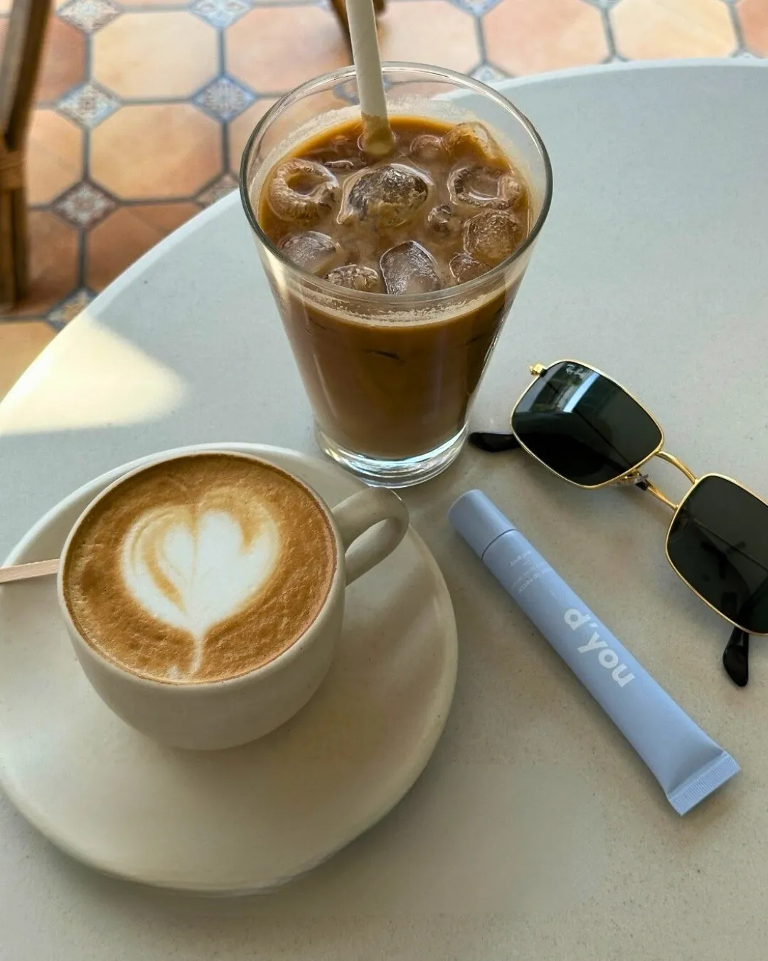 A d'you lip balm placed on a marble table with a cup of hot coffee and glass of iced chocolate and black sunnies