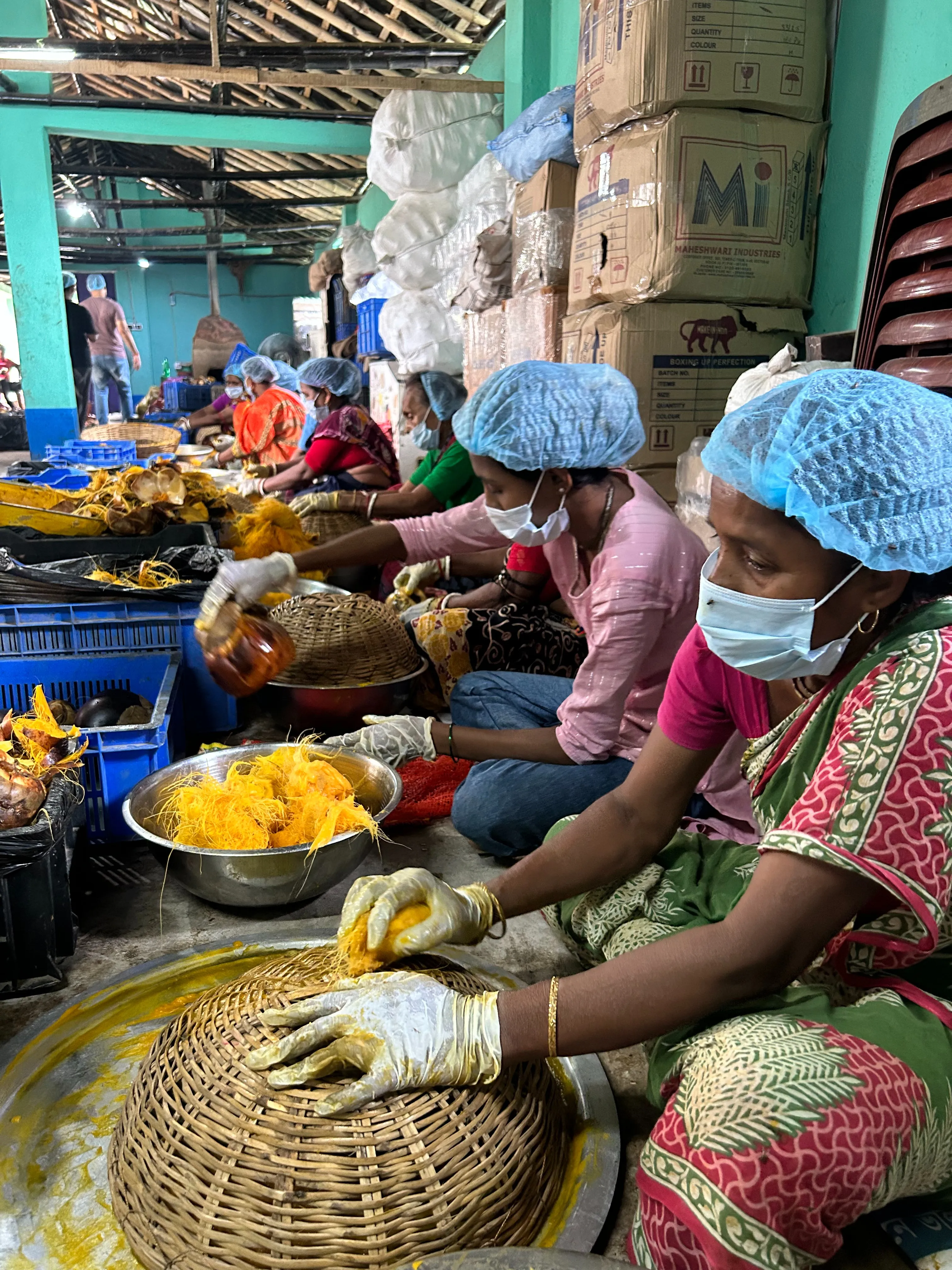 A group of female artisans in a processing facility wearing protective hairnets, masks, and gloves, traditionally extracting pulp from bright orange palm fruits using large wicker baskets
