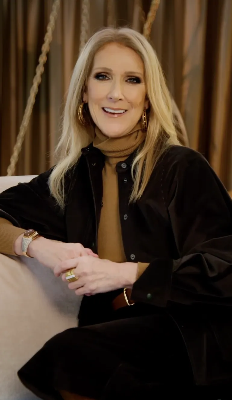 Celine Dion sitting and smiling, wearing a black jacket over a camel-colored turtleneck.