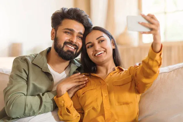 Loving indian couple sitting on sofa, embracing and taking selfie on  cellphone, looking at phone screen and smiling, free space, home interior.  Relationships concept - Stock Image - Everypixel