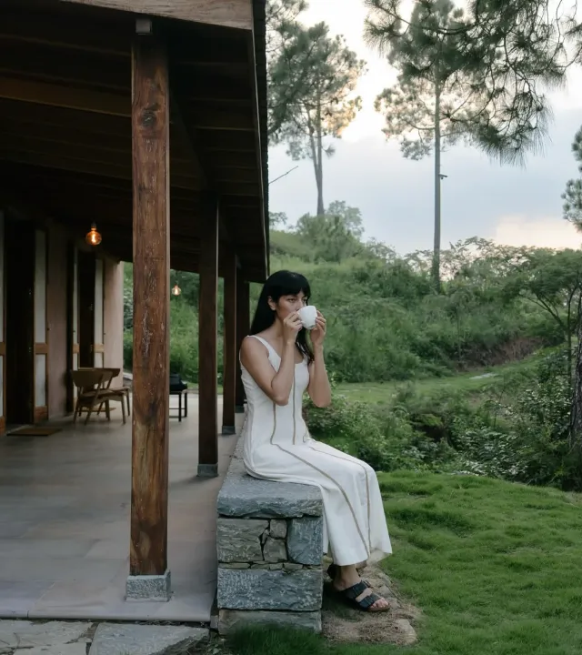 A woman dressed in white sips a cup of tea or coffee while sitting on a seating made using stone and a roof made using wood at Amaya, depicting the sustainable hotels India and impact of eco tourism India. The green hillside it overlooks represents how luxury resorts India also function as eco stay India