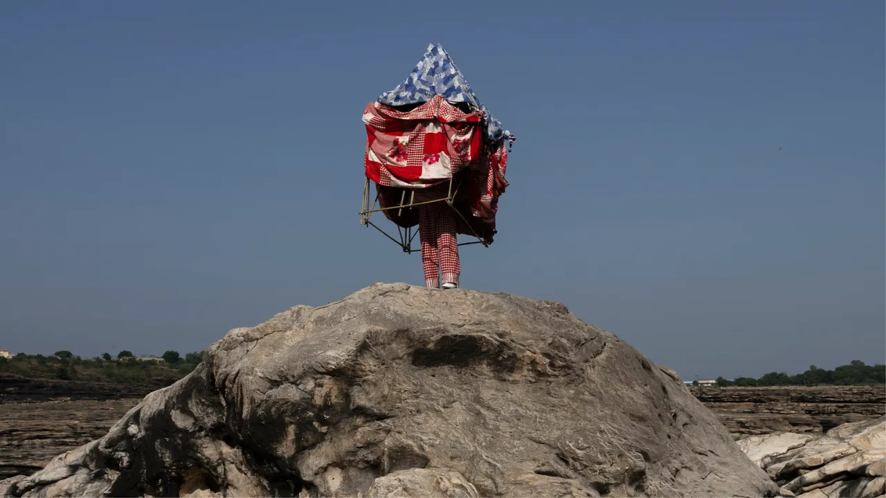  A figure standing on a rocky landscape wearing layered Péro textiles arranged into a sculptural, tent-like form, photographed as part of the Péro home line campaign 