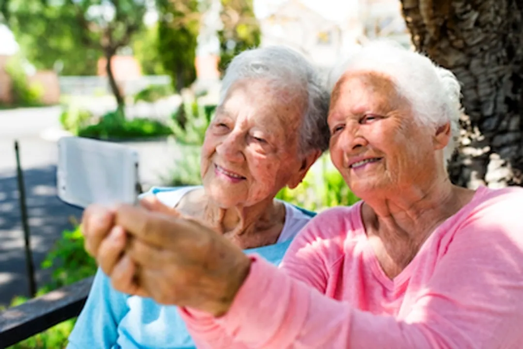  Two happy female senior citizens sitting outside posing together for a selfie. 