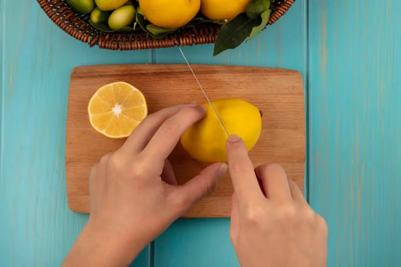 top-view-female-hands-cutting-fresh-lemon-wooden-kitchen-board-with-knife-with-fruits-such-as-kinkans-lemons-bucket-blue-wooden-surface_141793-81002