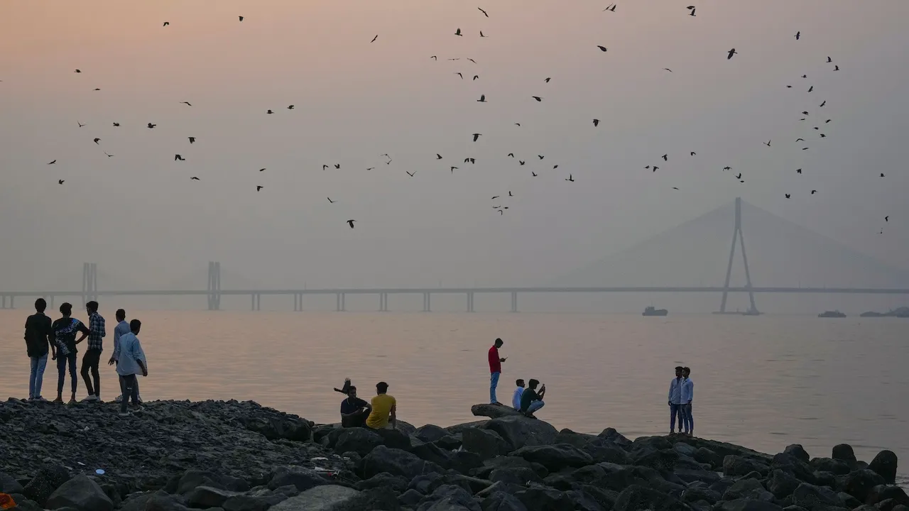 People sit along the shoreline at Dadar Beach as the Bandra-Worli Sea Link remains shrouded in smog, in Mumbai, Thursday, Nov. 27, 2025.