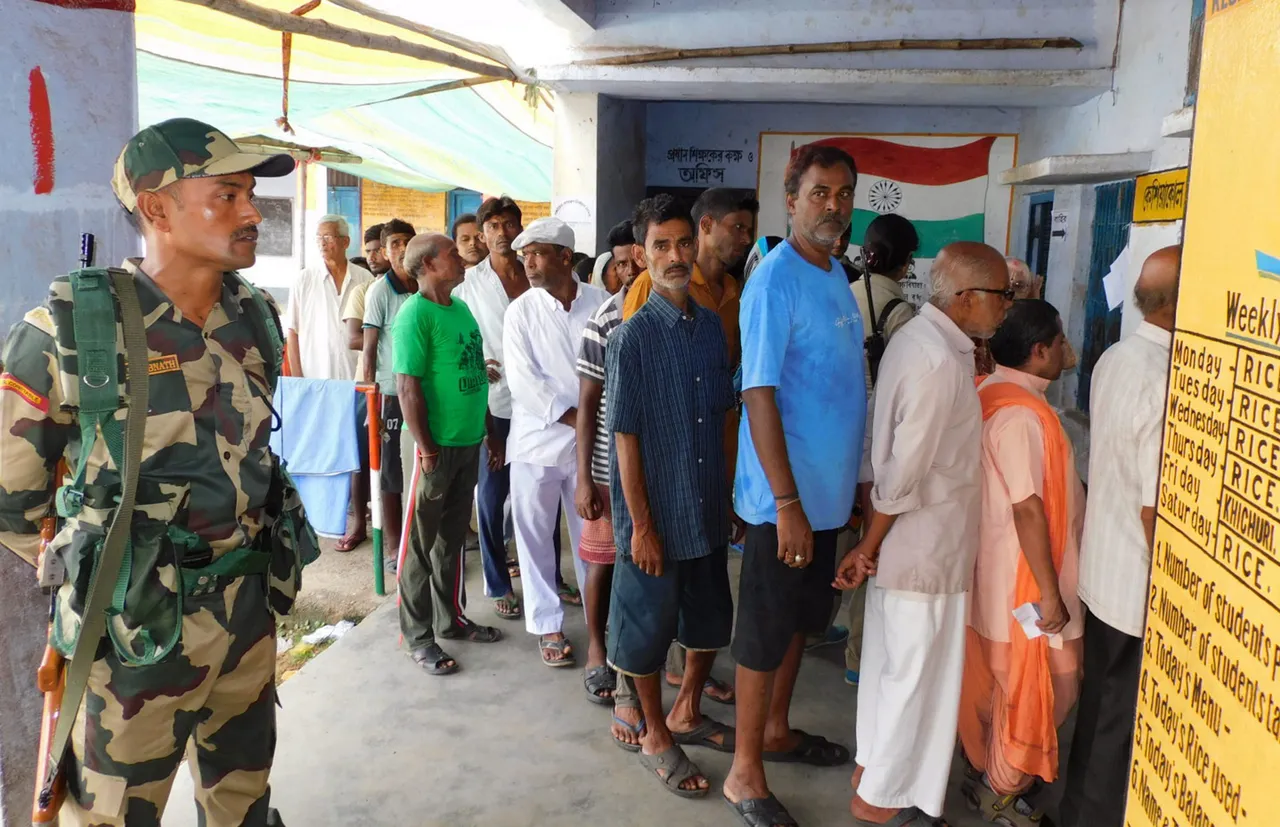 A security person stands guard as people stand in a queue to cast their votes for Panchayat elections at a polling station, in Bankura