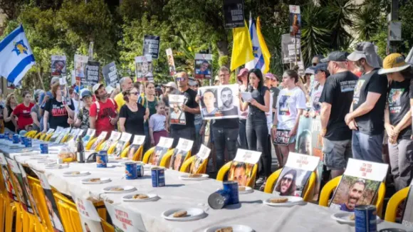 A landscape images of a table laid with cans and bread with the images of hostages taped to chairs. A group of people stand behind with flags, images and posters. One woman at the front speaks into a microphone
