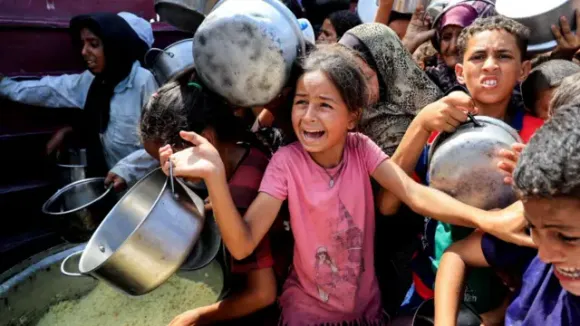 Palestinian children and women are crowded together, holding empty pans as they attemmpt to get food from an aid station providing meals