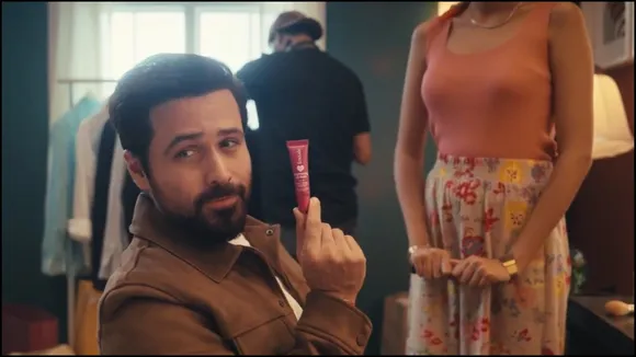 Actor Emraan Hashmi is shown smiling in a close-up shot, holding a tube of Foxtale's Brightening Lip Balm. His lips appear well-cared for.