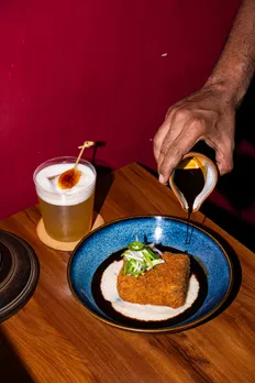 A blue plate with a cutlet being plated with a sauce being poured on it, while a drink is on the side