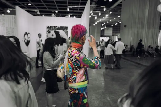 A black and white photograph of a person in colourful clothes and pink hair, depicting queer pride India and drag culture India, in a room full of people that's black and white to show queer safe spaces India and queer visibility India