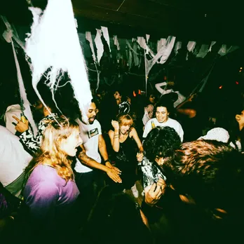 A room full of people crowding around a DJ as they sing and dance to show how the growing popularity of electronic music in India as well as the commercialisation of techno in India