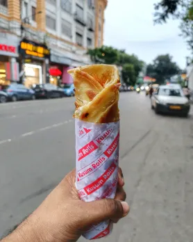A hand holds up a Kolkata Kati roll against the backdrop of a road to show where he mist-try Indian street food gets it's name, being the USP of street food Kolkata
