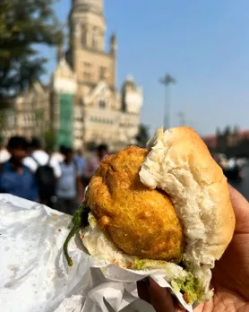 A hand holding up a vada pao against a background of Mumbai city to establish it as the core of street food Mumbai which is also one of the famous Indian snacks abroad.