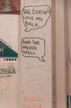 A sketch of two speech bubbles among other messages scribbled on a wall stating 'she doesn't love me back', with the 'and she never will' response, to depict how queer dating in India works after a relationship ends with respect to queer friendships after breakup