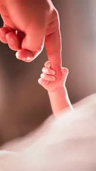A picture of an adult person's finger being held by a new born baby's hand to depict the rise of queer parenting in India and raising kids in queer families, despite incompatible surrogacy laws for queer couples India