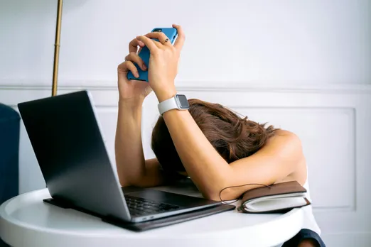 A picture of a woman putting her head down while holding her phone, in front of a laptop, to show how rage baiting in marketing is rampant, given that viral engagement and emotional marketing work well together in 2025