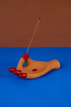 A terracotta-toned hand-shaped incense holder with red painted fingertips holding a lit incense stick against a brown and blue backdrop
