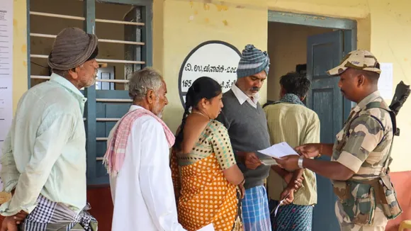 A security personnel checks voter ID as voters wait to cast votes at a polling station during the Channapatna Assembly by-poll, Karnataka