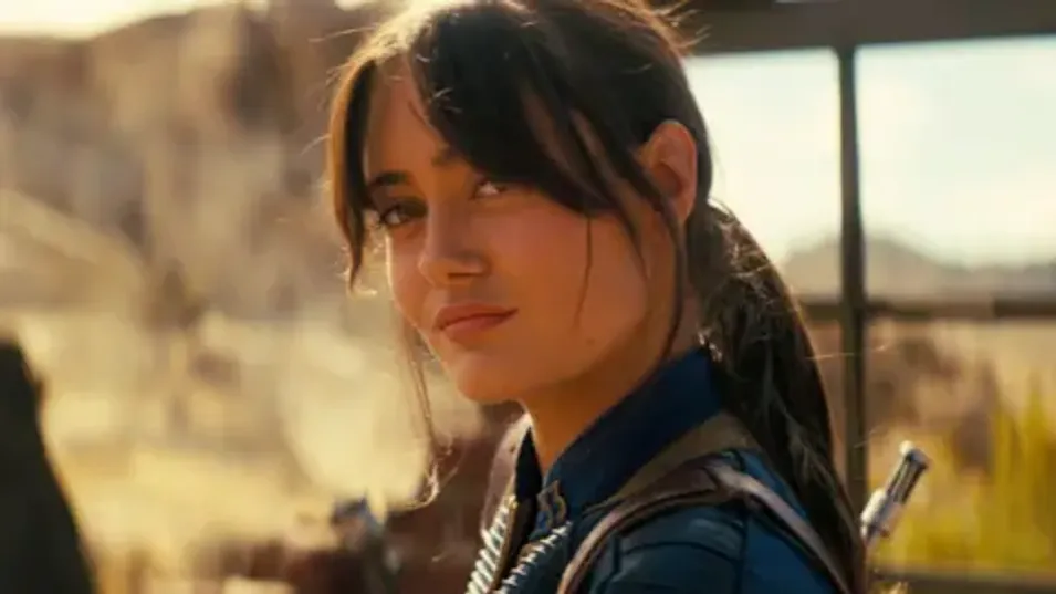 Prime Video A shot from Fallout shows Ella Purnell in her blue vault suit, hair tied back, looking quizzically at something in the distance. She's surrounded by desert and bathed in a dusky yellow sunlight. 
