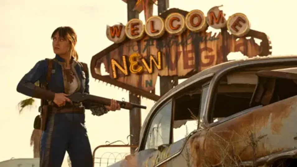 Prime Video Ella Purnell, dressed as character Lucy, stands next to a rusted, ruined car in front of a battered "Welcome to New Vegas" sign. She's holding a rifle as she looks off to the side, as if she's just spotted a sudden threat.