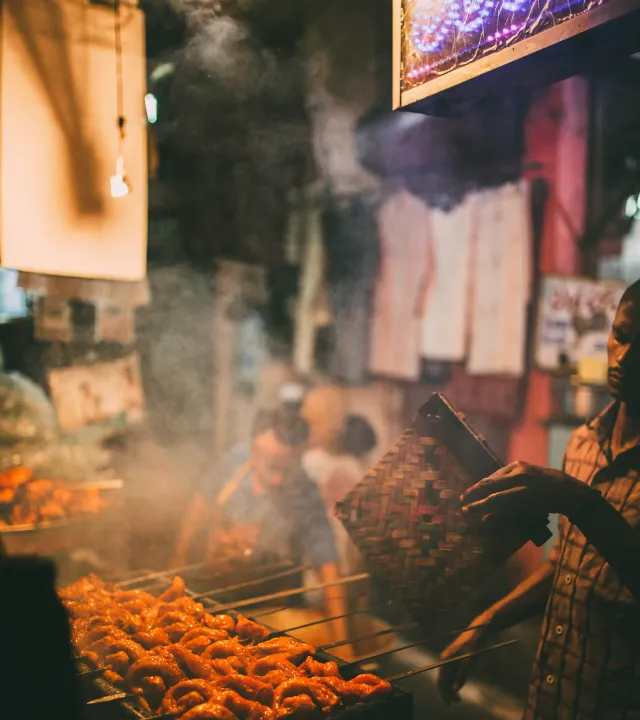 A man fans the coals of a tandoor on the roadside, as a kebabs sizzle on to depict kebabs, one of the best street food India, and famous Indian snacks that remain authentic.