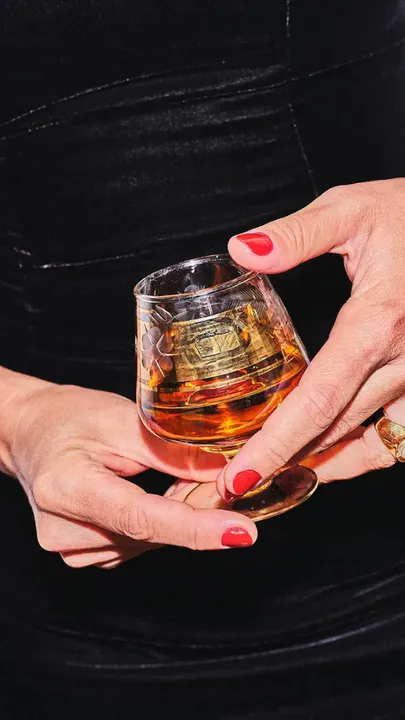 A picture of a woman in a black dress, holding glass of whiskey in her hands with red nail paint