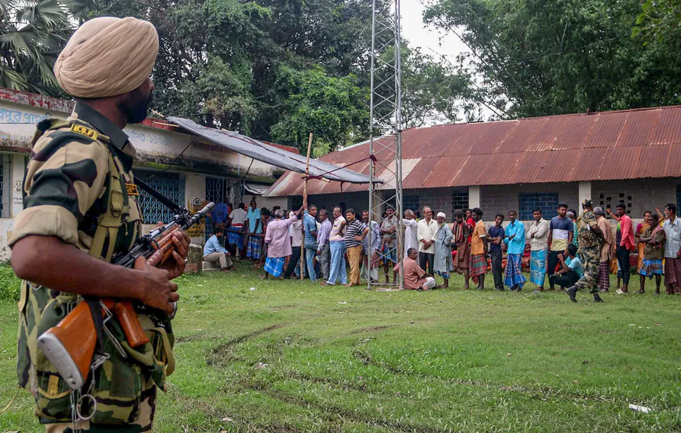A Border Security Force (BSF) personnel stand guard as people wait in queue to cast vote at a polling booth during Panchayat repolls, in South Dinajpur district, Monday